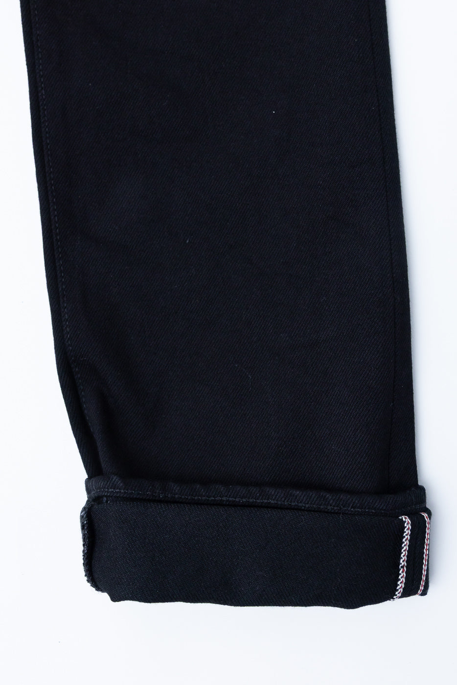 Close-up of black selvedge denim pant leg, front view, showing cuffed hem with red & white stitching detail