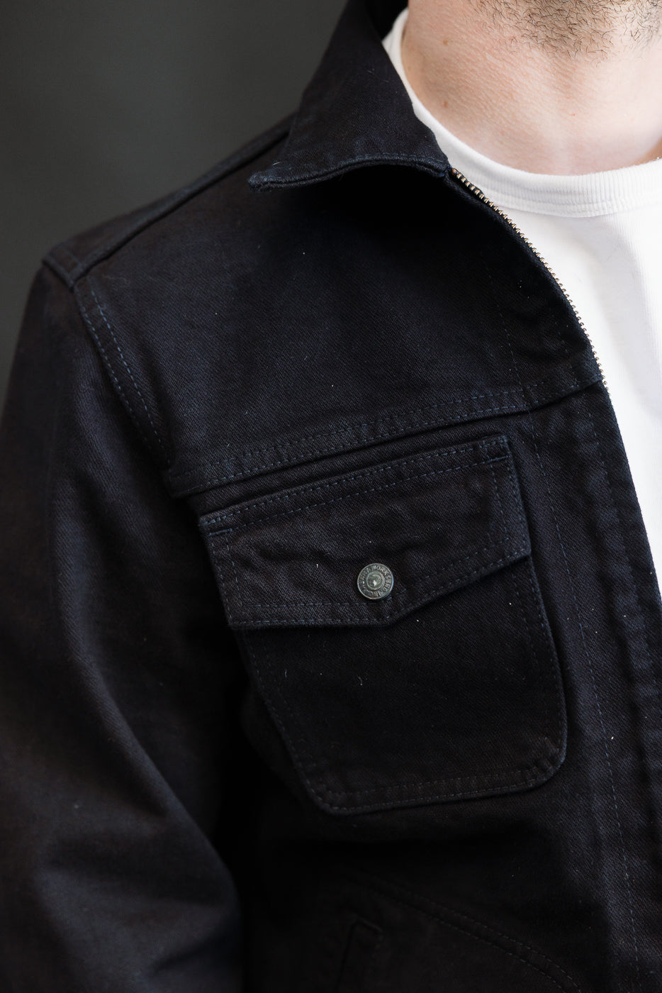 Close-up of the IHJ-123-OD - 14oz Broken Twill Selvedge Denim Work Jacket, showcasing its dark indigo overdyed black fabric, metal snap pocket, and folded collar over a white T-shirt.