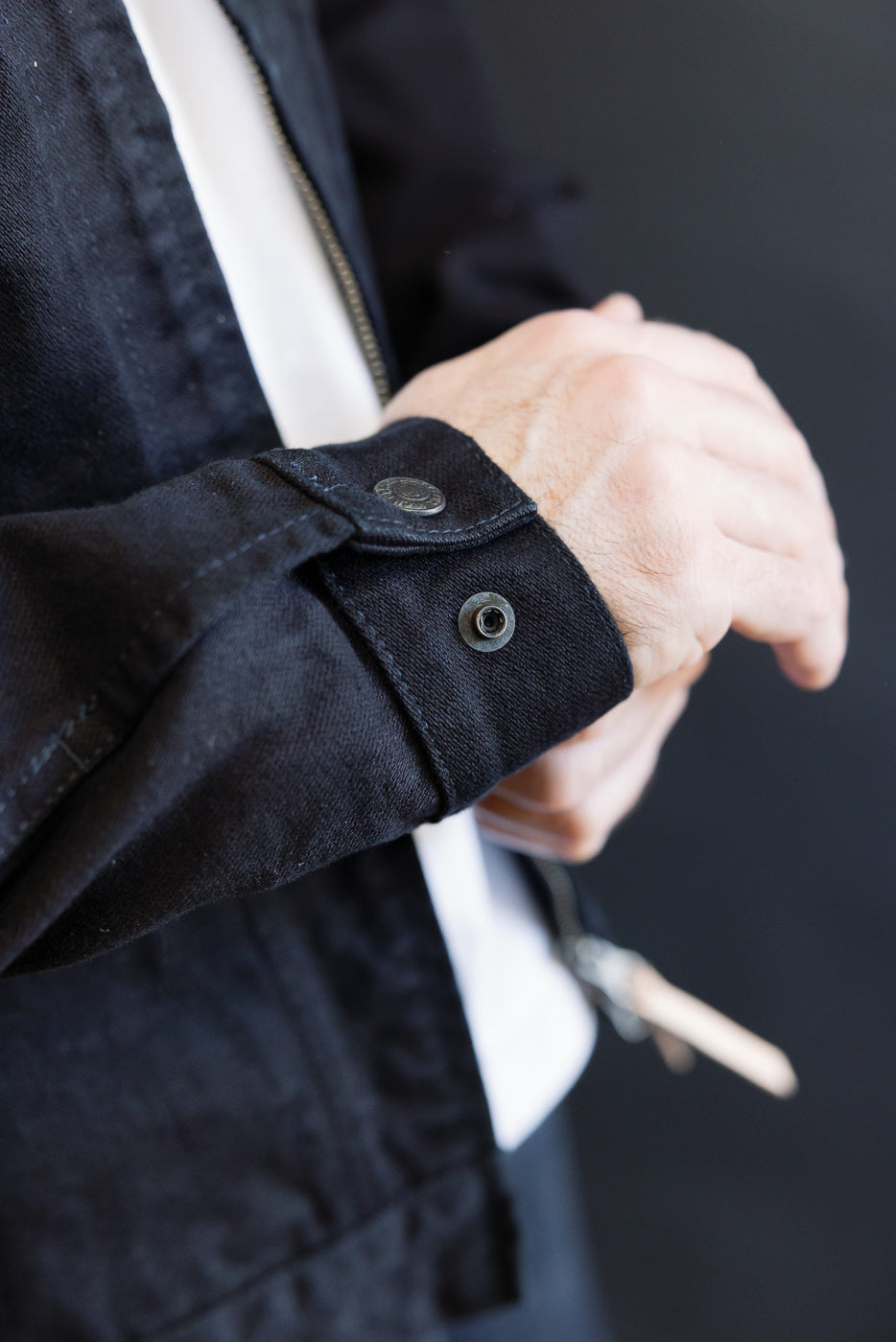 Close-up of a person adjusting the cuff of the IHJ-123-OD - 14oz Broken Twill Selvedge Denim Work Jacket, revealing its black snap button and part of the white shirt underneath.