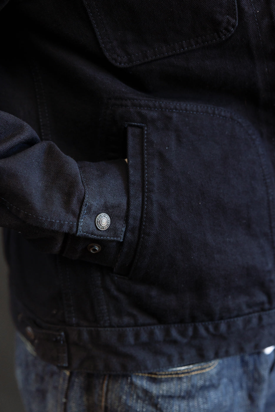 Close-up of the IHJ-123-OD - 14oz Broken Twill Selvedge Denim Work Jacket in Indigo Overdyed Black, showcasing the metal snap button cuff and hand tucked into a pocket, paired with blue jeans.