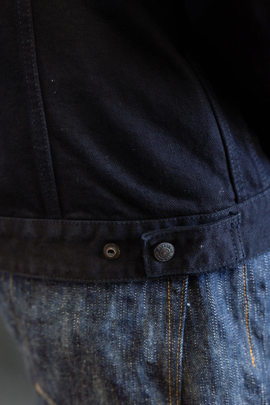 Close-up of the IHJ-123-OD - 14oz Broken Twill Selvedge Denim Work Jacket, highlighting the adjustable snap-button tab on the black hem, resting against blue jeans, showcasing meticulous craftsmanship.