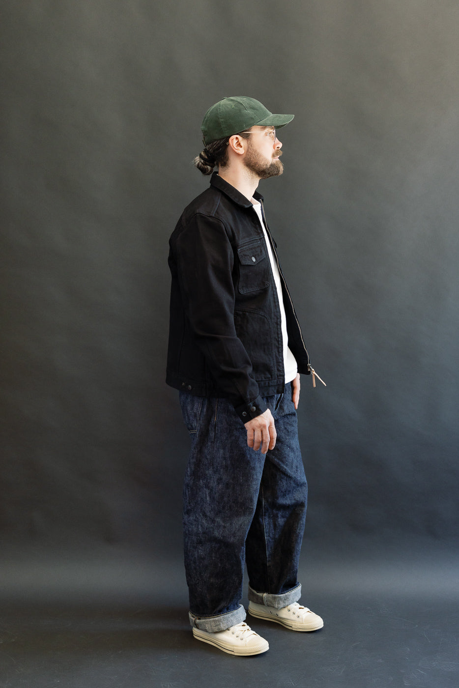 Man in profile wearing IHJ-123-OD, a 14oz Broken Twill Selvedge Denim Work Jacket, layered over a white T-shirt, paired with loose indigo jeans and canvas sneakers. Model's hair under green cap.