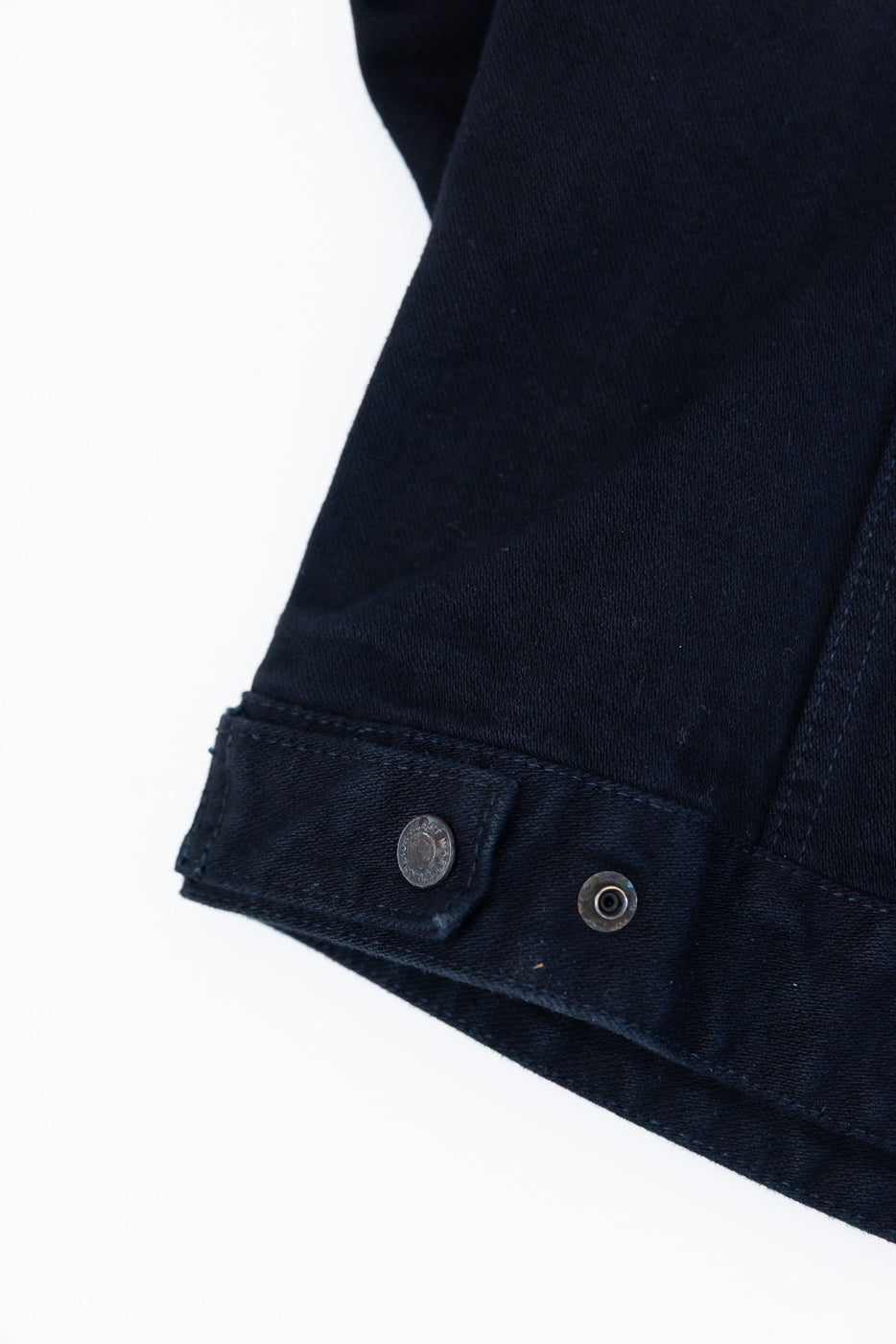 Close-up of the IHJ-123-OD - 14oz Broken Twill Selvedge Denim Work Jacket's dark-navy hem, showcasing the stitched adjustment tab and metal snap-button hardware on a white background.