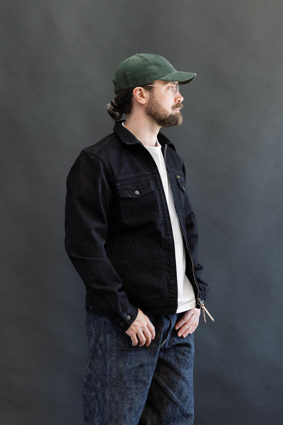 A man models the IHJ-123-OD - 14oz Broken Twill Selvedge Denim Work Jacket - Indigo Overdyed Black, featuring a black zip-front design with flap pockets, highlighting its craftsmanship and heritage style.