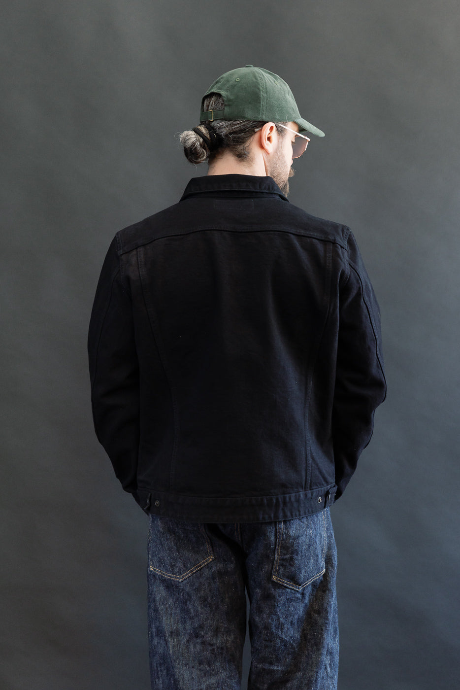 Person in a dark denim jacket, IHJ-123-OD - 14oz Broken Twill Selvedge Denim Work Jacket, paired with blue jeans, showcasing a casual utilitarian style, with cap and sunglasses, in studio.