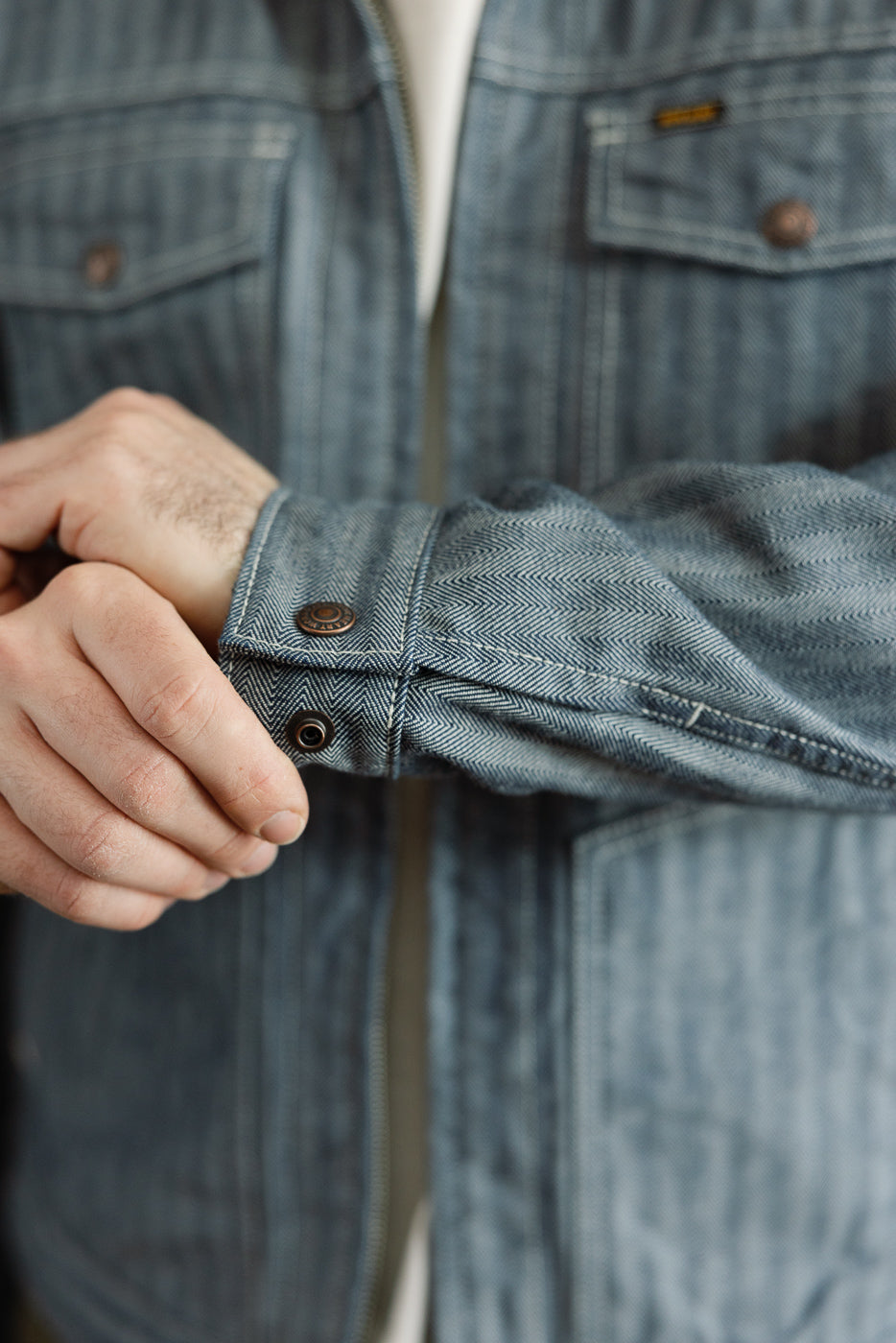 Close-up of male model, 5'9, 165lbs, athletic build, adjusting indigo herringbone work jacket cuff, showing front fabric texture and button detail.