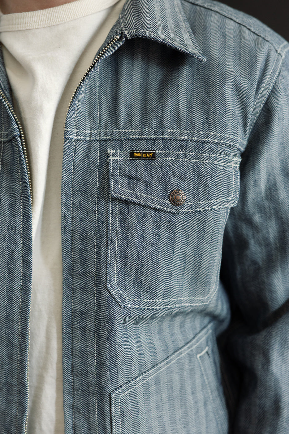 Close-up of front left chest and pocket of indigo herringbone work jacket on male model, highlighting metal button, visible tag, pointed collar, and textured fabric; model is male, 5'9