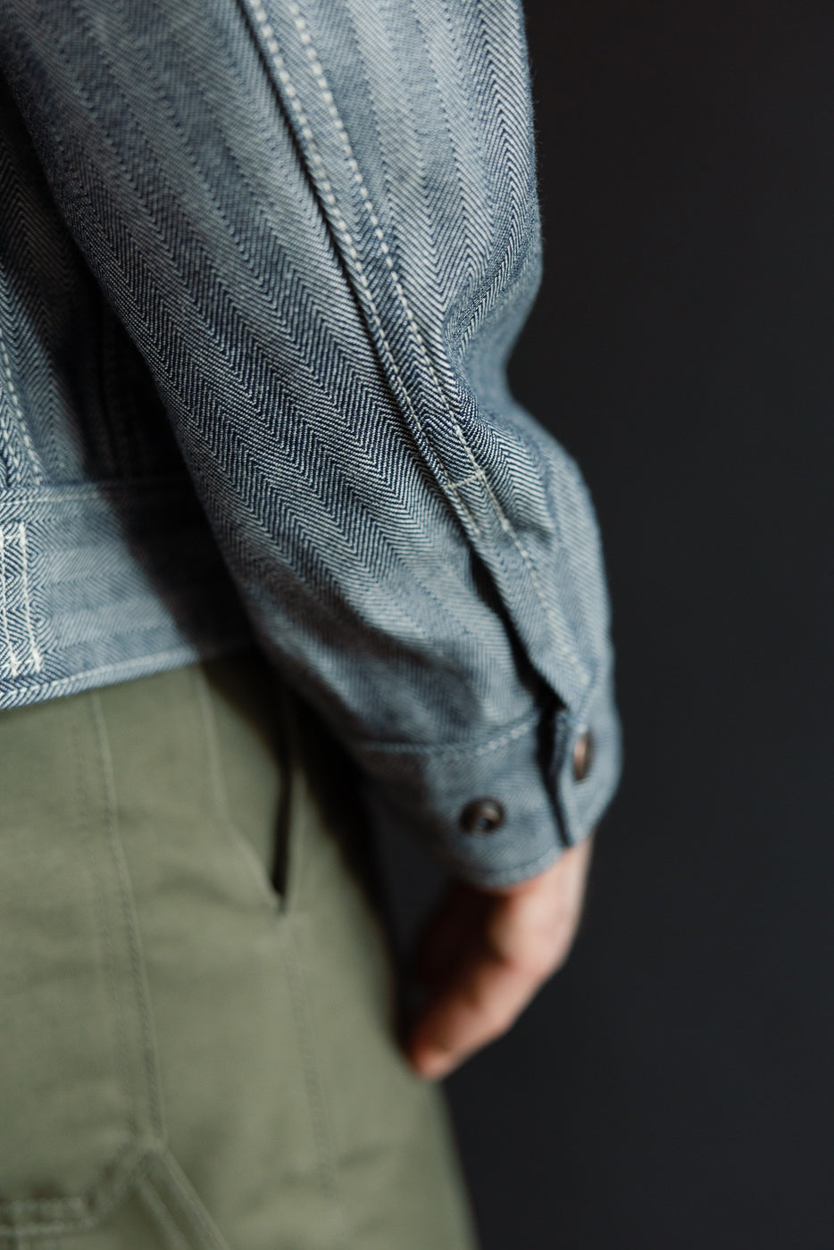 Close-up of the right sleeve cuff and side seam on a male model wearing a herringbone work jacket, highlighting indigo fabric texture and button details. Model is 5'9