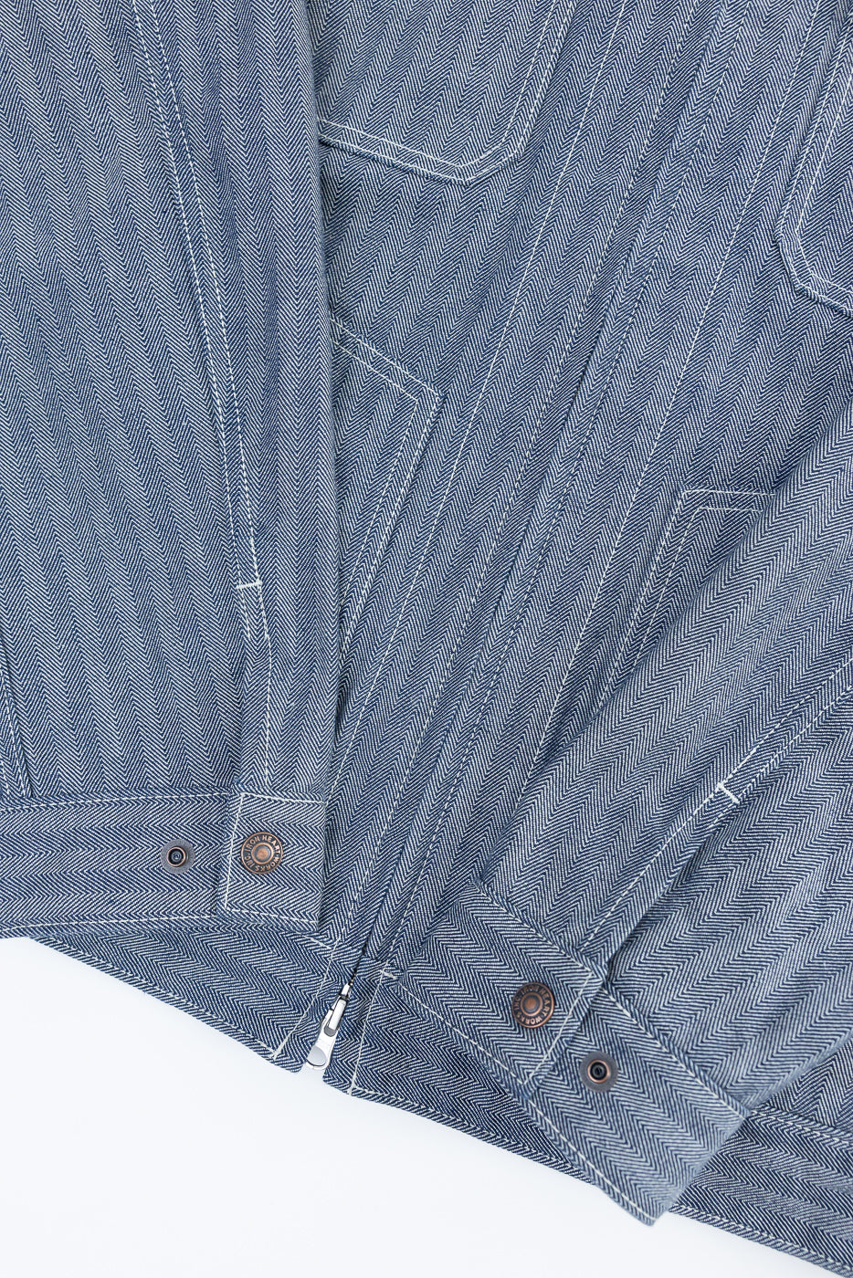 Close-up of indigo herringbone work jacket front hem, showing snap buttons, zipper, and fabric texture.