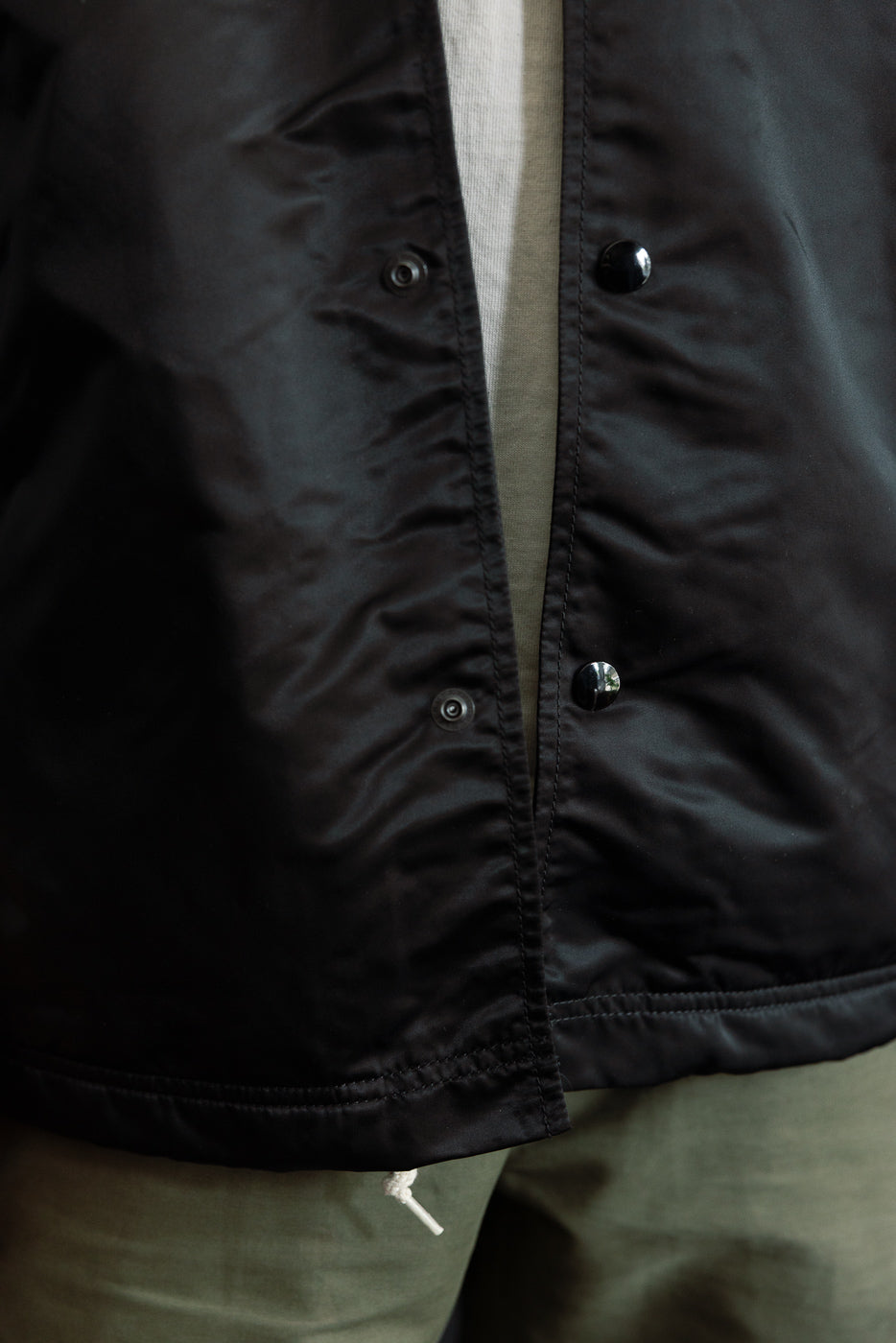 Close-up front view of nylon black coach jacket snap buttons and hem details on male model.
