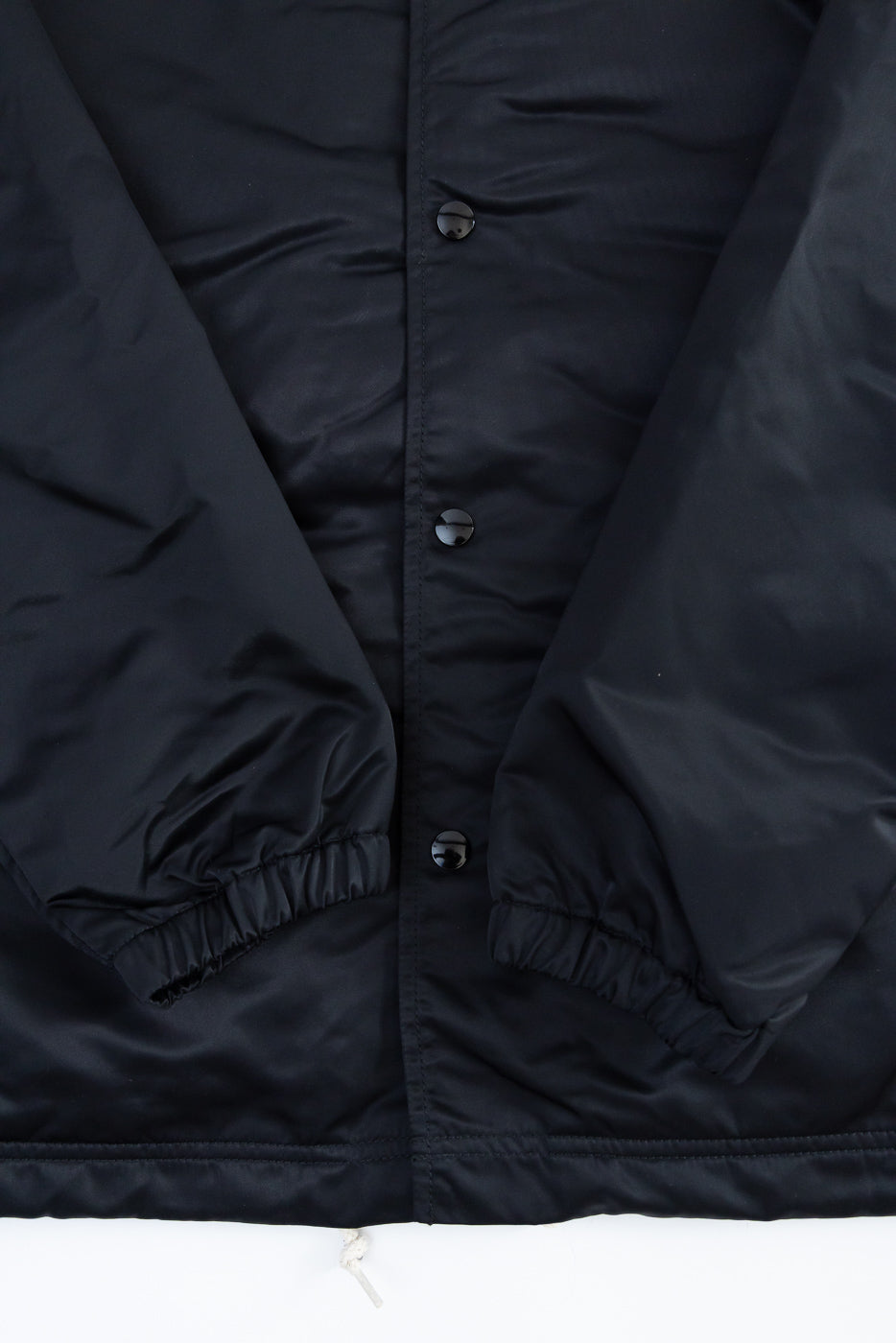 Close-up of front midsection of black nylon coach jacket showing snap buttons, elastic cuffs, and hem.