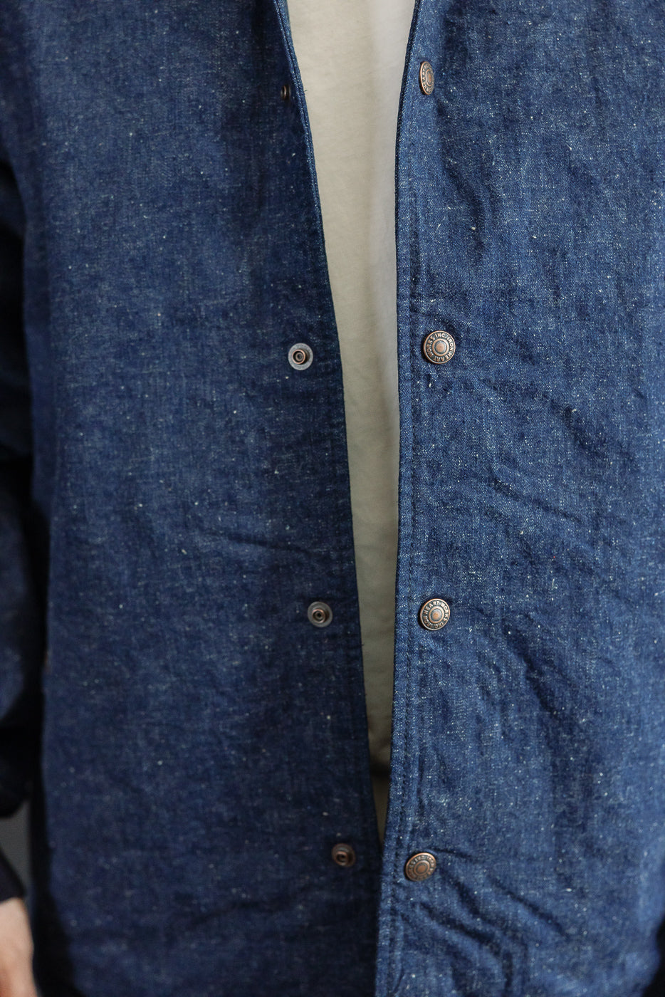 Close-up front view of men's indigo denim shirt, showing button placket, texture, and brass buttons on athletic build model