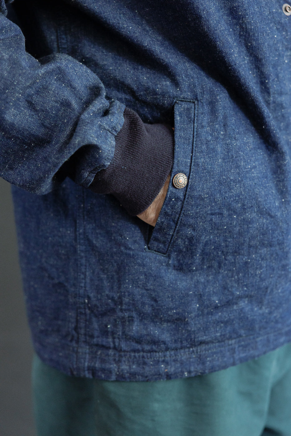 Close-up of male model's side pocket on indigo denim shirt, showcasing button detail and fabric texture.