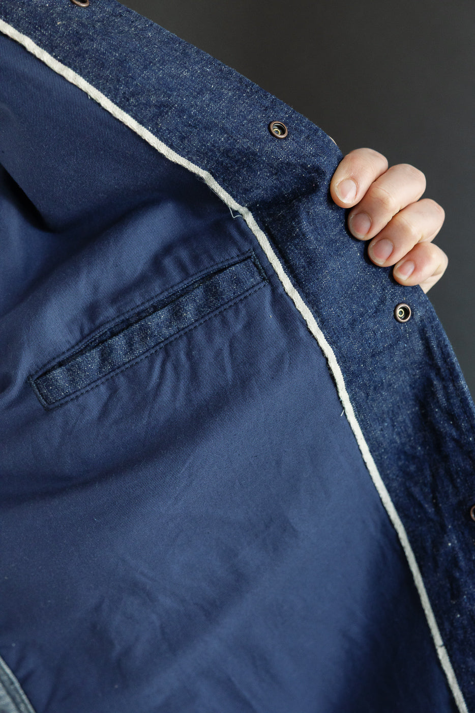 Close-up of indigo denim jacket front, showcasing button details and white piping, held by hand.