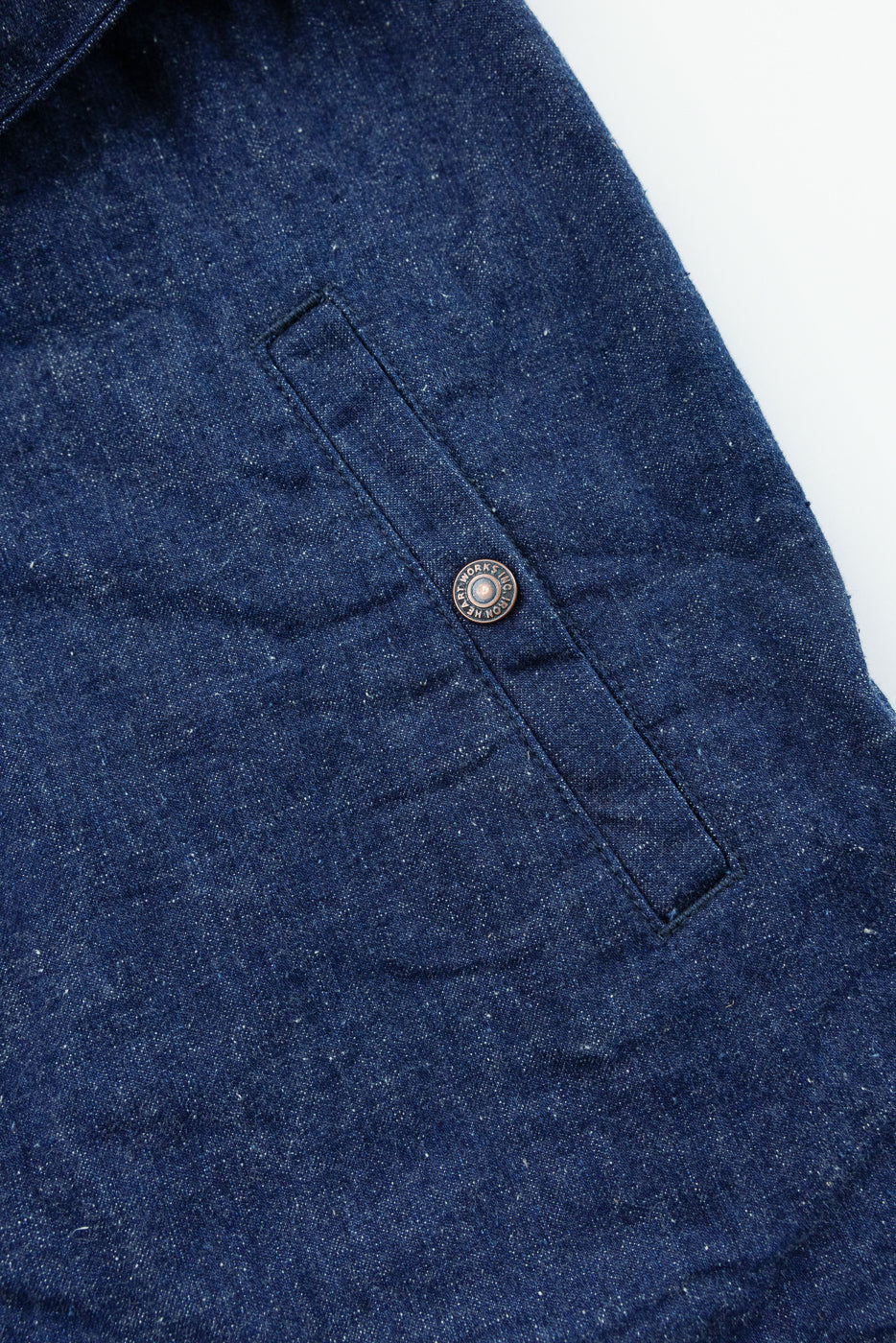 Close-up front view of dark denim shirt pocket with branded button, showcasing fabric texture and stitching details.