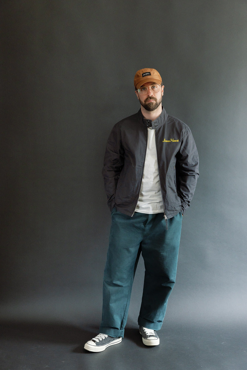 Full-body front view of male model in dark jacket, teal pants, and cap, showcasing casual apparel.