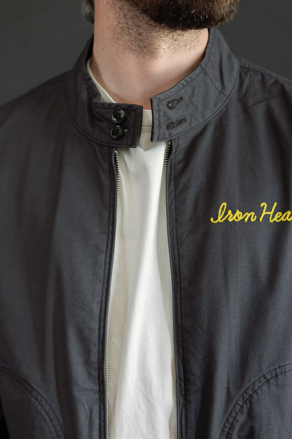Close-up front view of male model wearing dark jacket with yellow 'Iron Head' embroidery and buttoned collar.