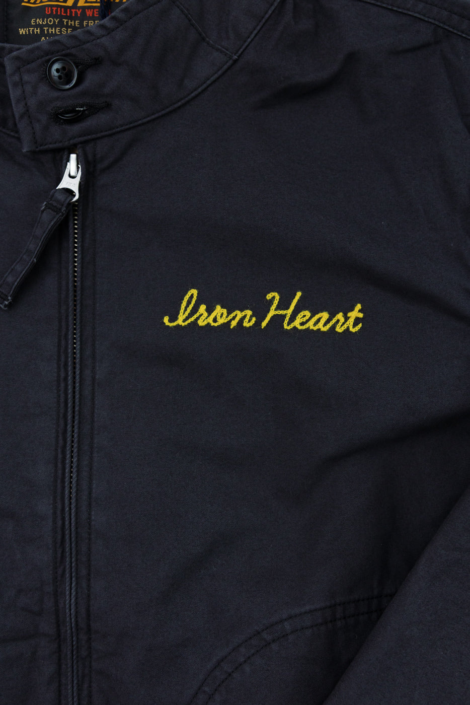 Close-up front view of black jacket with yellow 'Iron Heart' embroidery, zipper, and buttoned collar detail