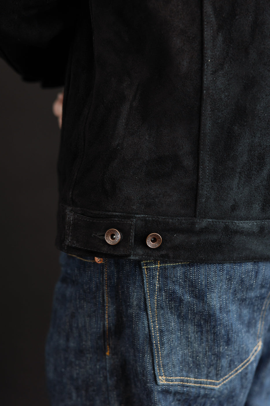 Close-up side view of black suede jacket hem with copper snap buttons, worn by male model, paired with raw denim jeans.
