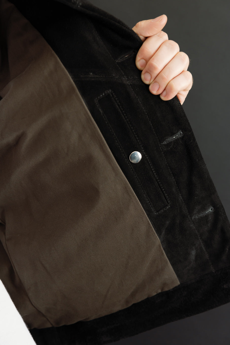Close-up of black suede jacket front, showing silver button, pocket detail, and brown lining, held by male model.