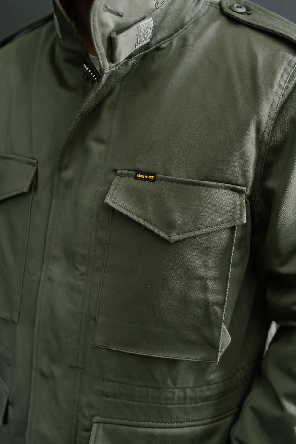 Close-up front view of Iron Heart olive sateen M65 field jacket chest pockets, collar, and epaulet on male model, 5'9