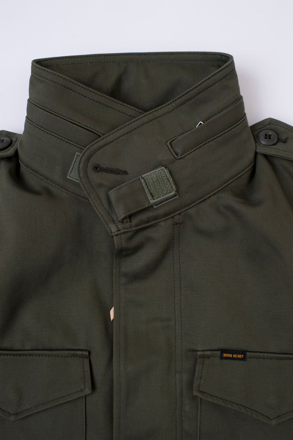Close-up of Iron Heart olive green M-65 field jacket front, showing stand collar, button epaulettes, pocket flaps, and chest logo tag in sateen fabric.