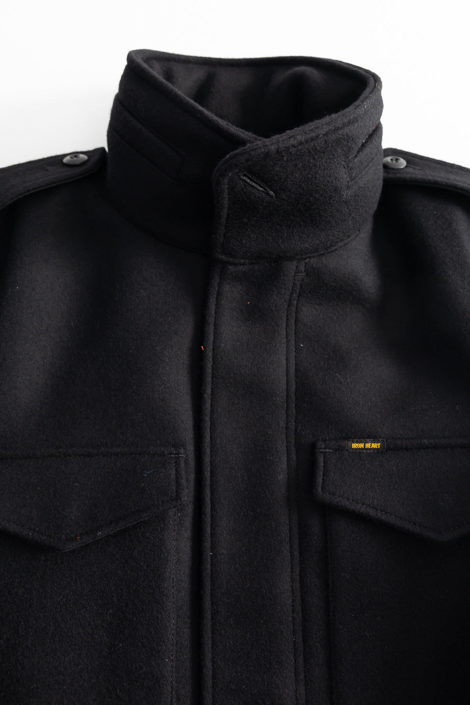 Close-up front view of Iron Heart IHJ-49-BLK 21oz melton wool M65 field jacket collar and chest pockets, showing button shoulder epaulets and Iron Heart fabric tag, for Dant contemporary casual apparel.
