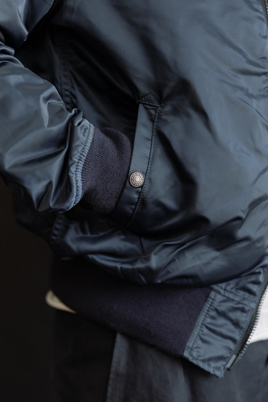 Close-up of male model wearing navy nylon jacket, showing right front pocket, snap button, ribbed cuff, and hem. Model is 5'9