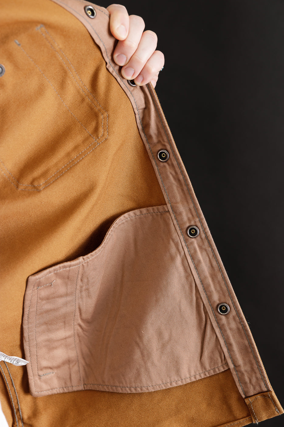 Close-up of male model holding open brown work shirt, showing interior stitching, pocket, and snap button details, inside front panel visible.