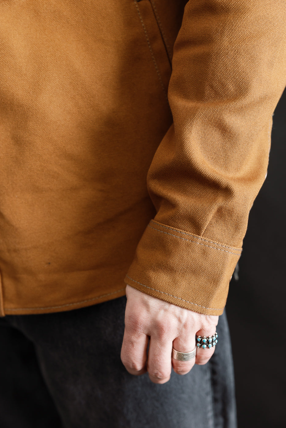 Close-up of male model's right sleeve and cuff of brown duck work shirt, showing fabric texture and stitching. Model is 5'9