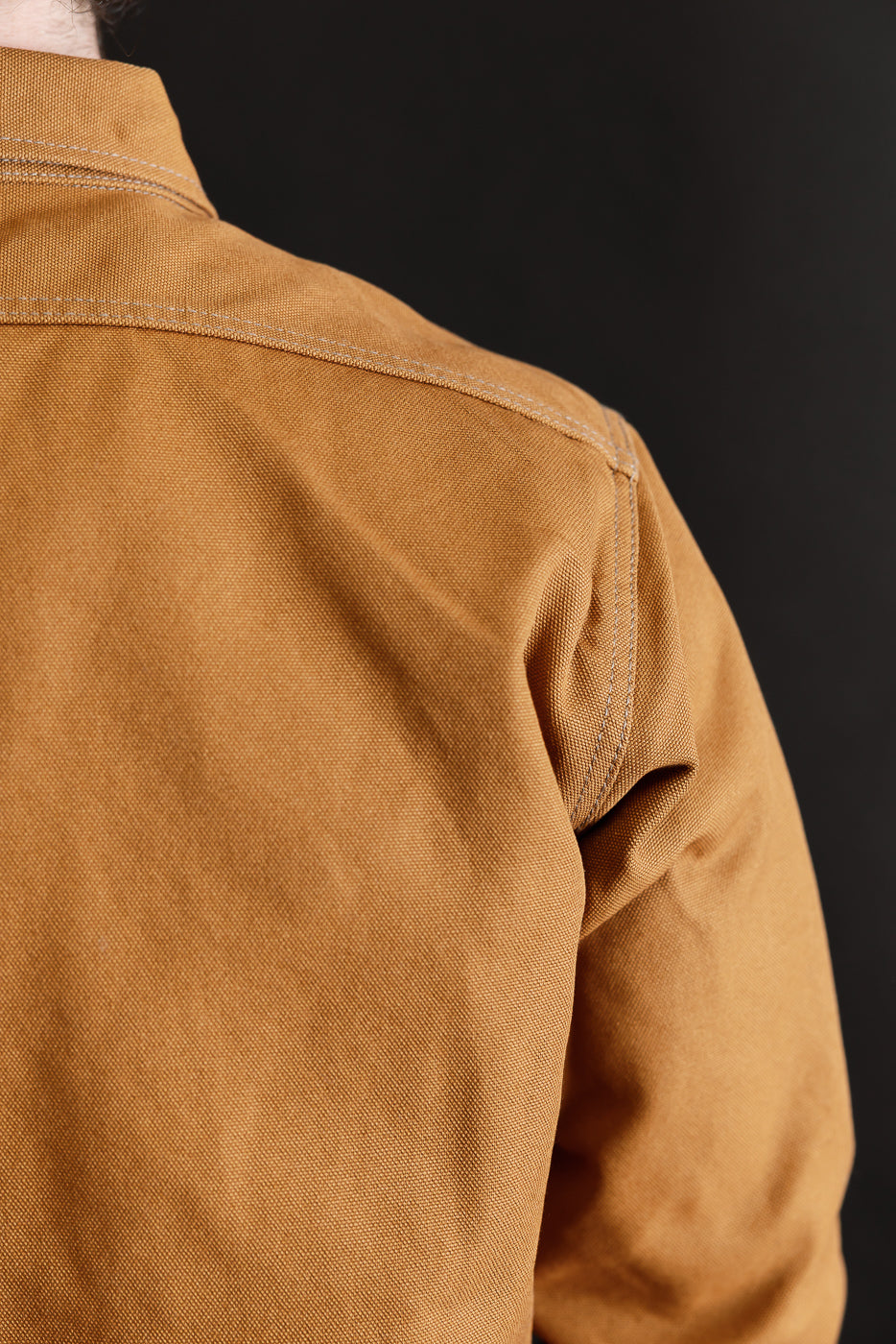 Close-up of men's brown work shirt, rear shoulder and upper sleeve, highlighting thick duck canvas texture and stitching. Model is male, 5'9