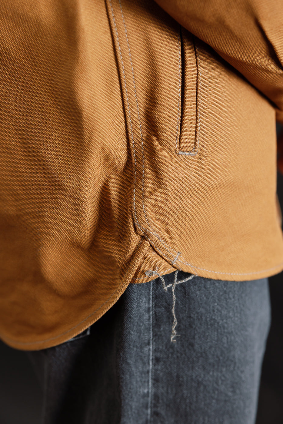 Close-up of lower side hem and pocket seam of brown work shirt, showing fabric texture and stitching detail.