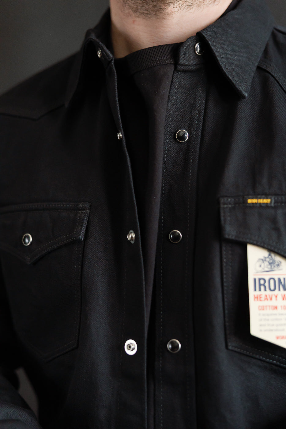 Close-up front of Iron Heart black selvedge denim western shirt on male model, showing snap buttons, chest pockets, and fabric detail. Model is 5'9