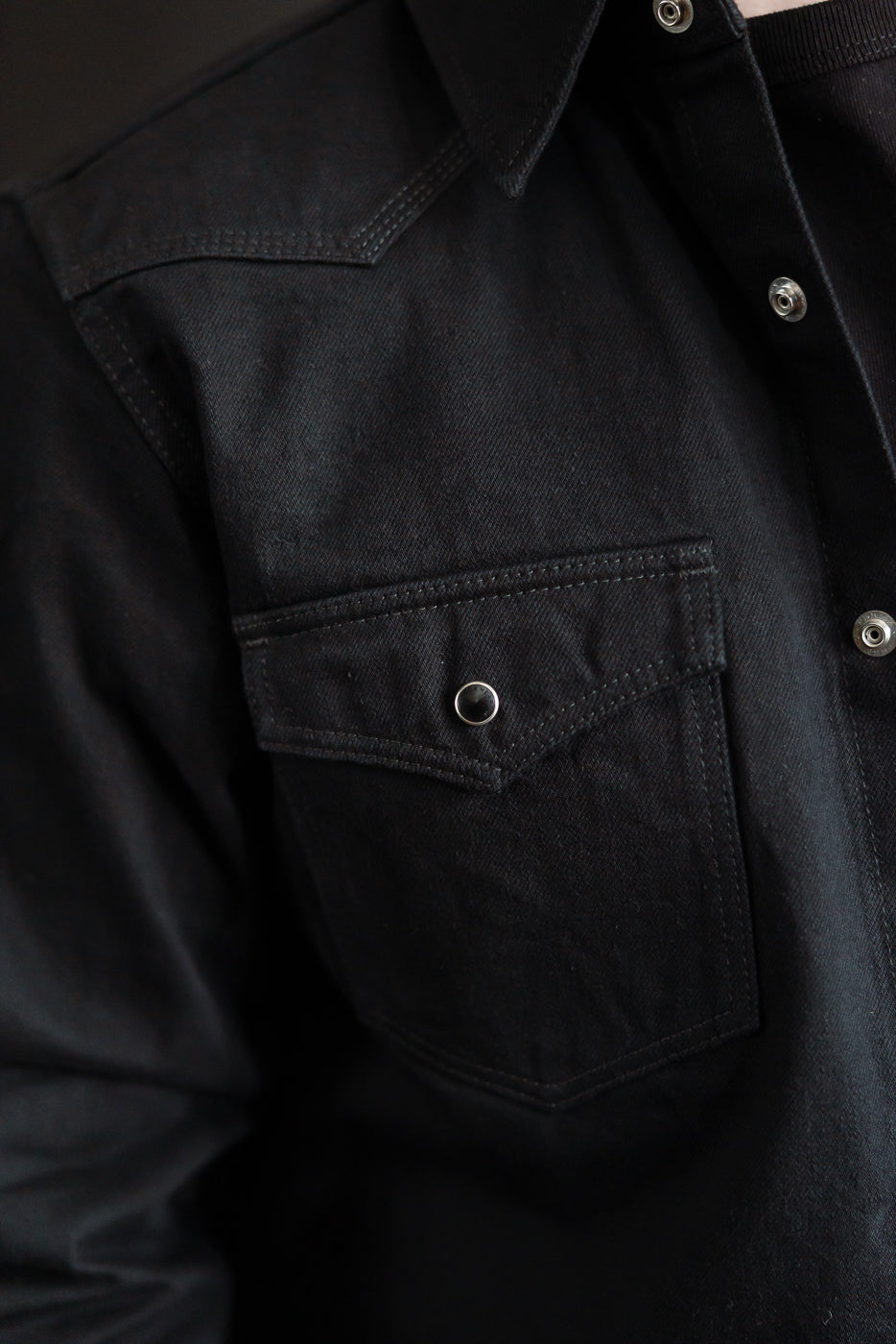 Close-up of front left chest pocket, snap button, and collar on black selvedge denim western shirt.