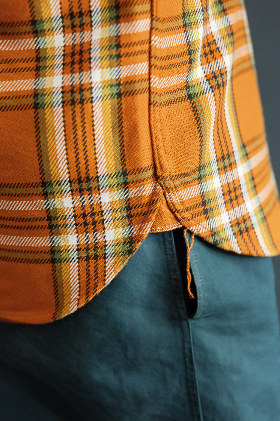 Close-up of orange plaid shirt hem, front view, worn by male model, paired with teal pants