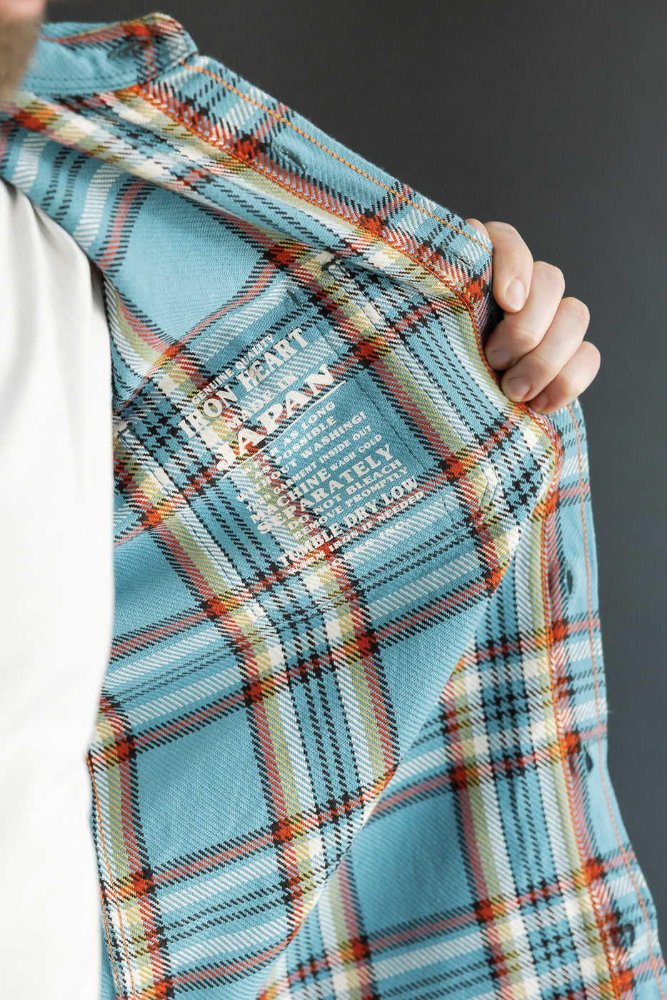 Close-up of male model's hand holding plaid shirt sleeve, showcasing tag details and fabric texture.