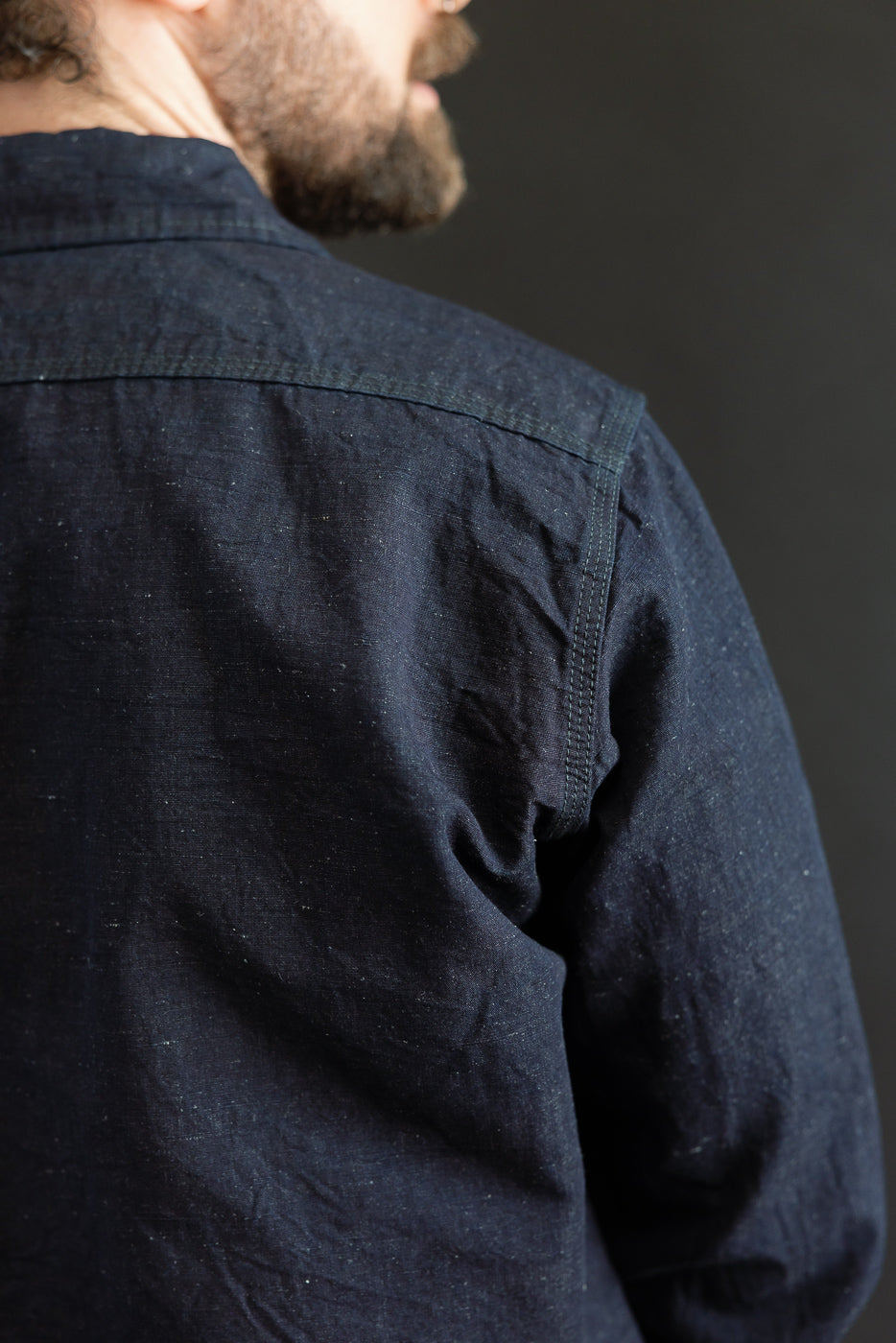 Close-up of male model's upper back in indigo chambray work shirt, showing back yoke and shoulder stitching. Model is 5'9