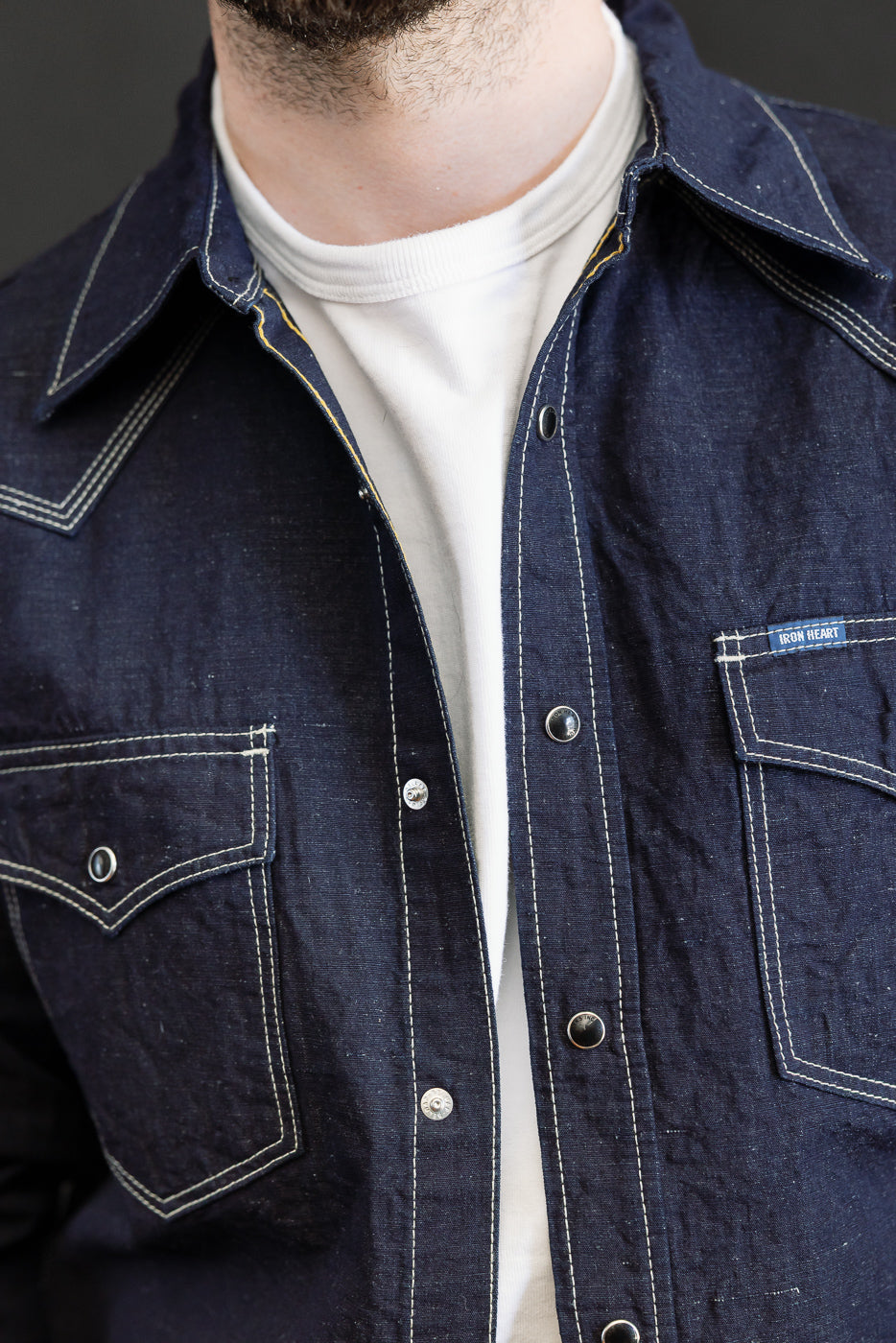 Close-up of the IHSH-444-IND - 7oz Cotton Linen Chambray Western Shirt in indigo, showcasing white contrast stitching, snap-button closures, and flap pockets with an IRON HEART label.
