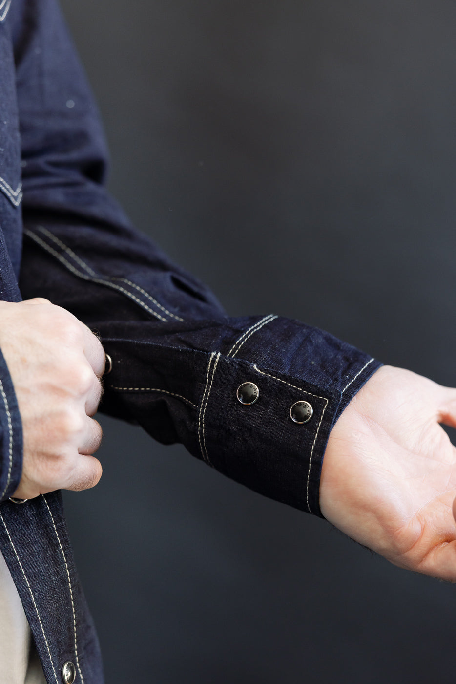 Close-up of IHSH-444-IND - 7oz Cotton Linen Chambray Western Shirt, showcasing the dark indigo denim jacket's cuff with contrasting white stitching and metal snap buttons, emphasizing fabric detail.