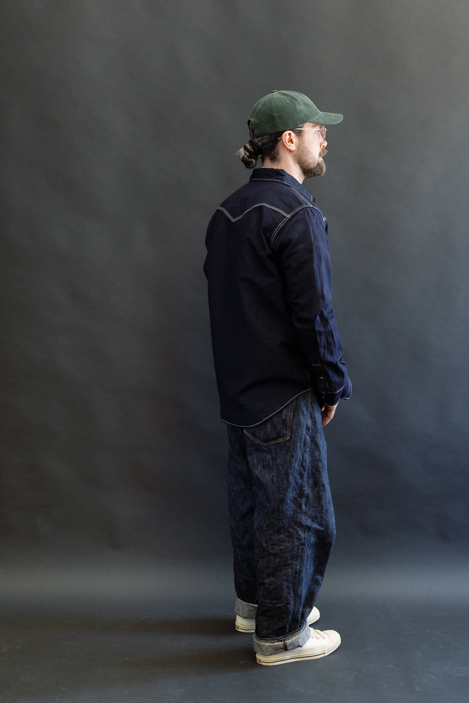 A man models the IHSH-444-IND - 7oz Cotton Linen Chambray Western Shirt, showcasing its indigo hue, contrast stitching, and western-style yokes, paired with cuffed jeans and sneakers.