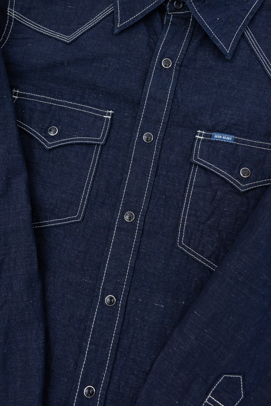 Close-up of the IHSH-444-IND - 7oz Cotton Linen Chambray Western Shirt in indigo, featuring white contrast stitching, snap-button closures, and two chest flap pockets with an Iron Heart woven label.
