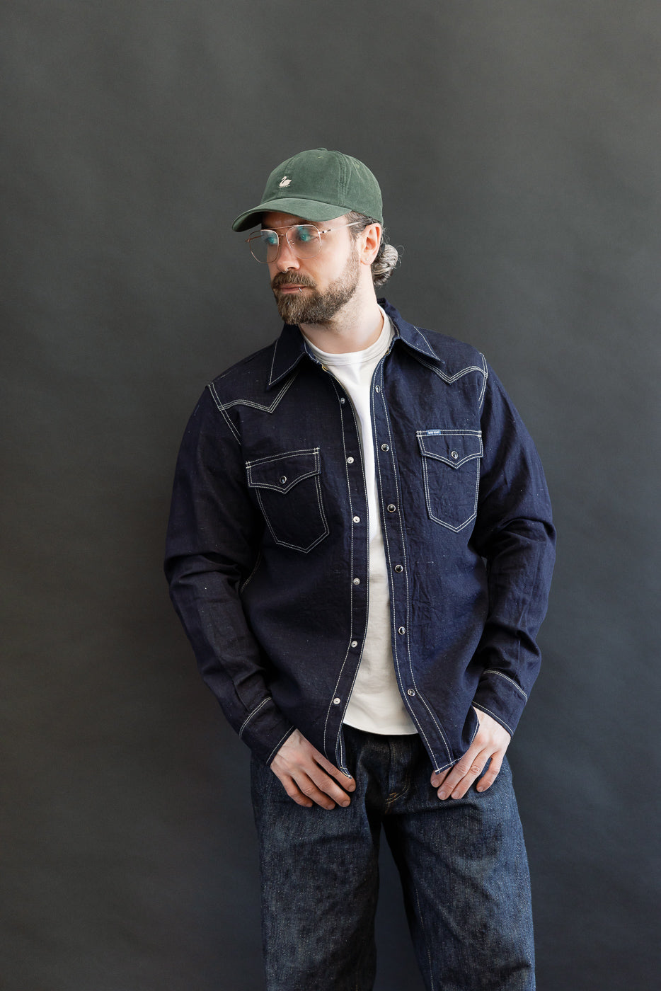Man wearing IHSH-444-IND - 7oz Cotton Linen Chambray Western Shirt in indigo, featuring snap fastenings, flap-down pockets, and contrast stitching, styled with matching dark blue jeans and a forest-green baseball cap.
