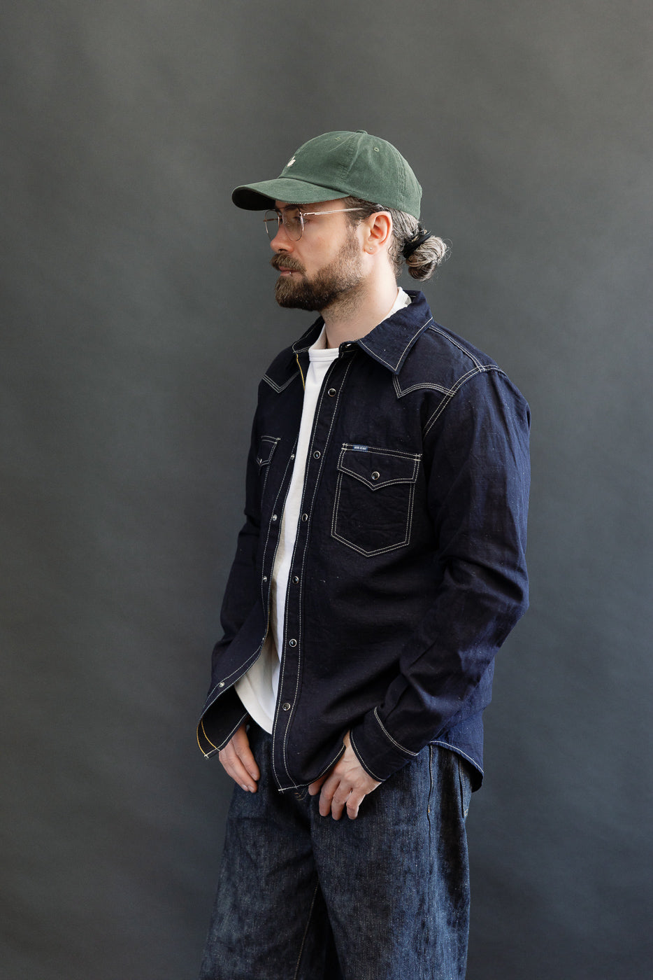 Man in profile wearing the IHSH-444-IND - 7oz Cotton Linen Chambray Western Shirt, indigo color, styled open over a white T-shirt, paired with denim trousers and a forest-green baseball cap.