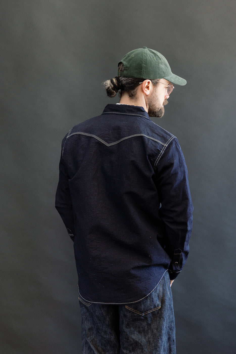Back view of a male model wearing the IHSH-444-IND, a 7oz Cotton Linen Chambray Western Shirt in indigo, featuring contrast stitching, paired with matching denim jeans and a green baseball cap.