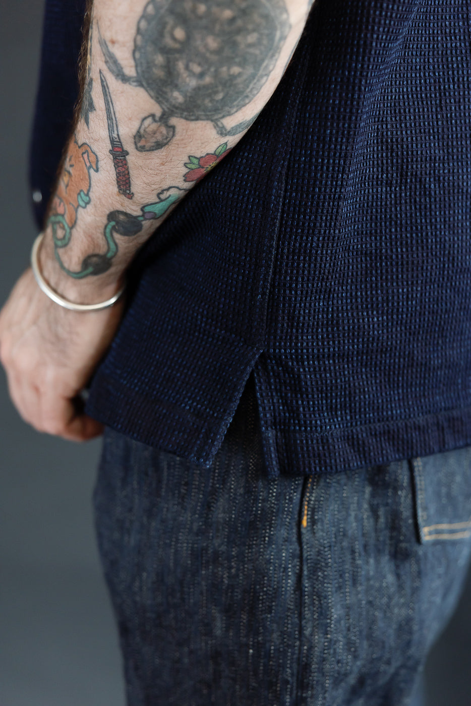 Close-up of male model's side, showcasing navy textured shirt hem and denim pants, tattooed arm visible