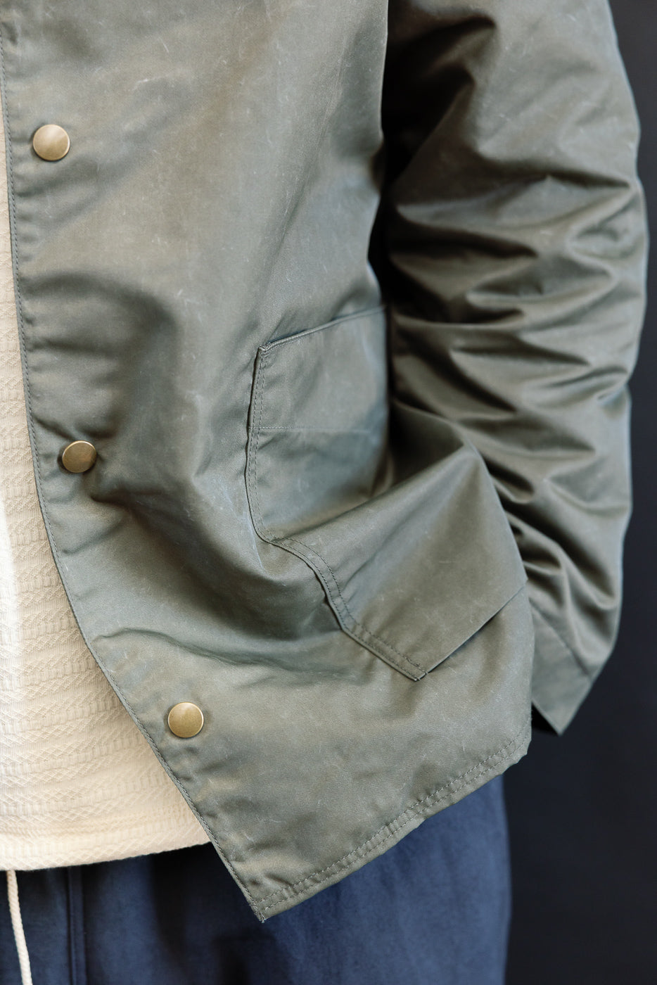 Close-up of male model wearing olive collarless jacket, front left pocket and snap buttons visible