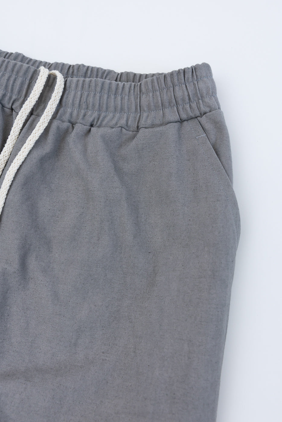 Close-up of front left side of gray canvas pants, showing waistband, drawstring, and pocket detail.