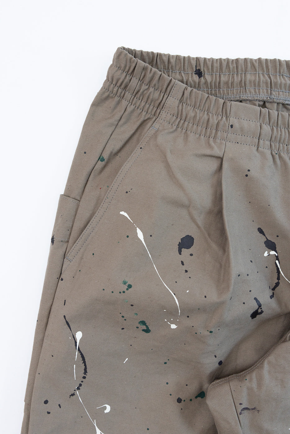 Close-up front view of tan paint-splattered casual trousers showing elastic waistband and pocket.