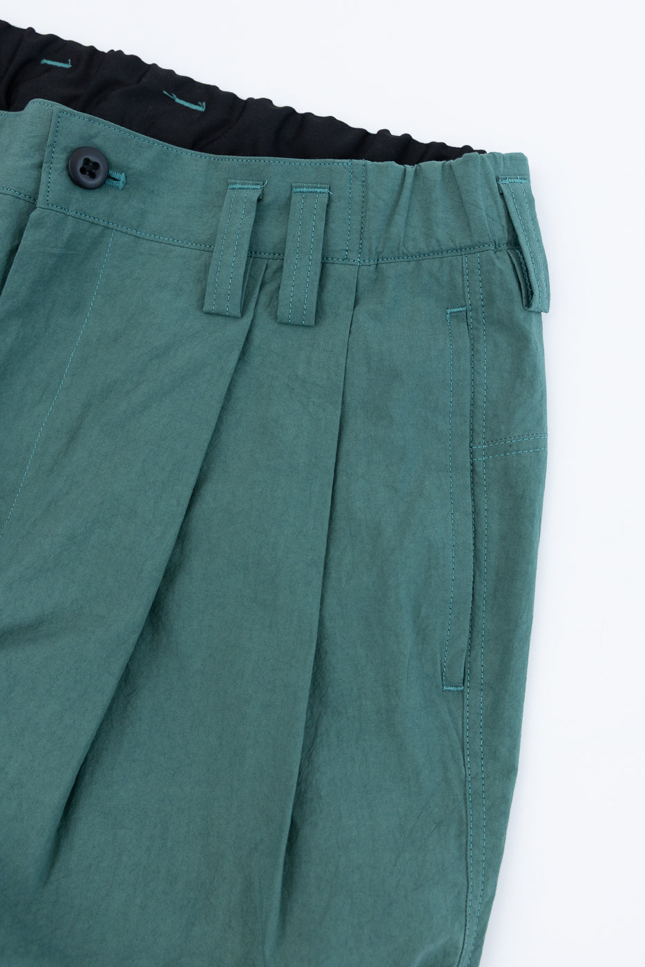 Close-up front view of teal pleated pants with black button, contrast stitching, and elastic waistband detail