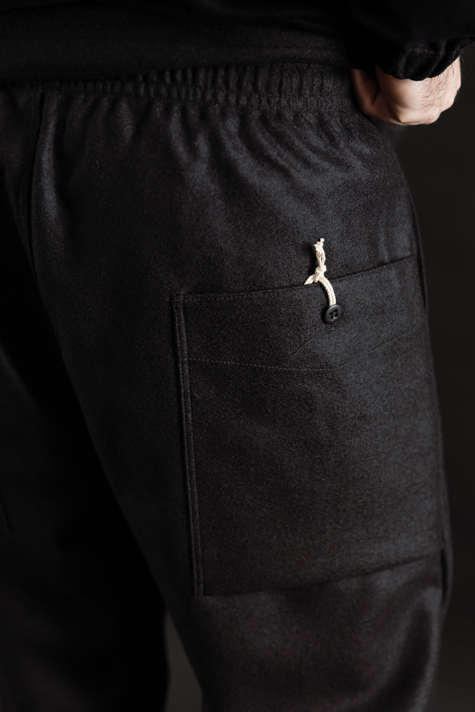 Close-up of male model's back right side, showing black wool moleskin trousers with patch pocket, button and drawstring detail. Model is 5'9